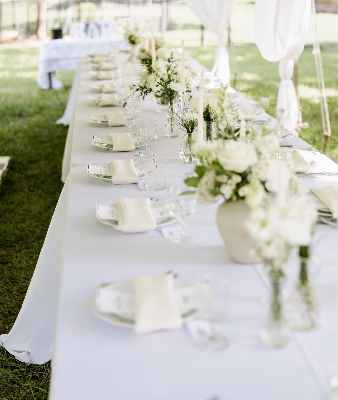 A long reception table set outdoors with white linens, folded napkins and white flowers in bud vases