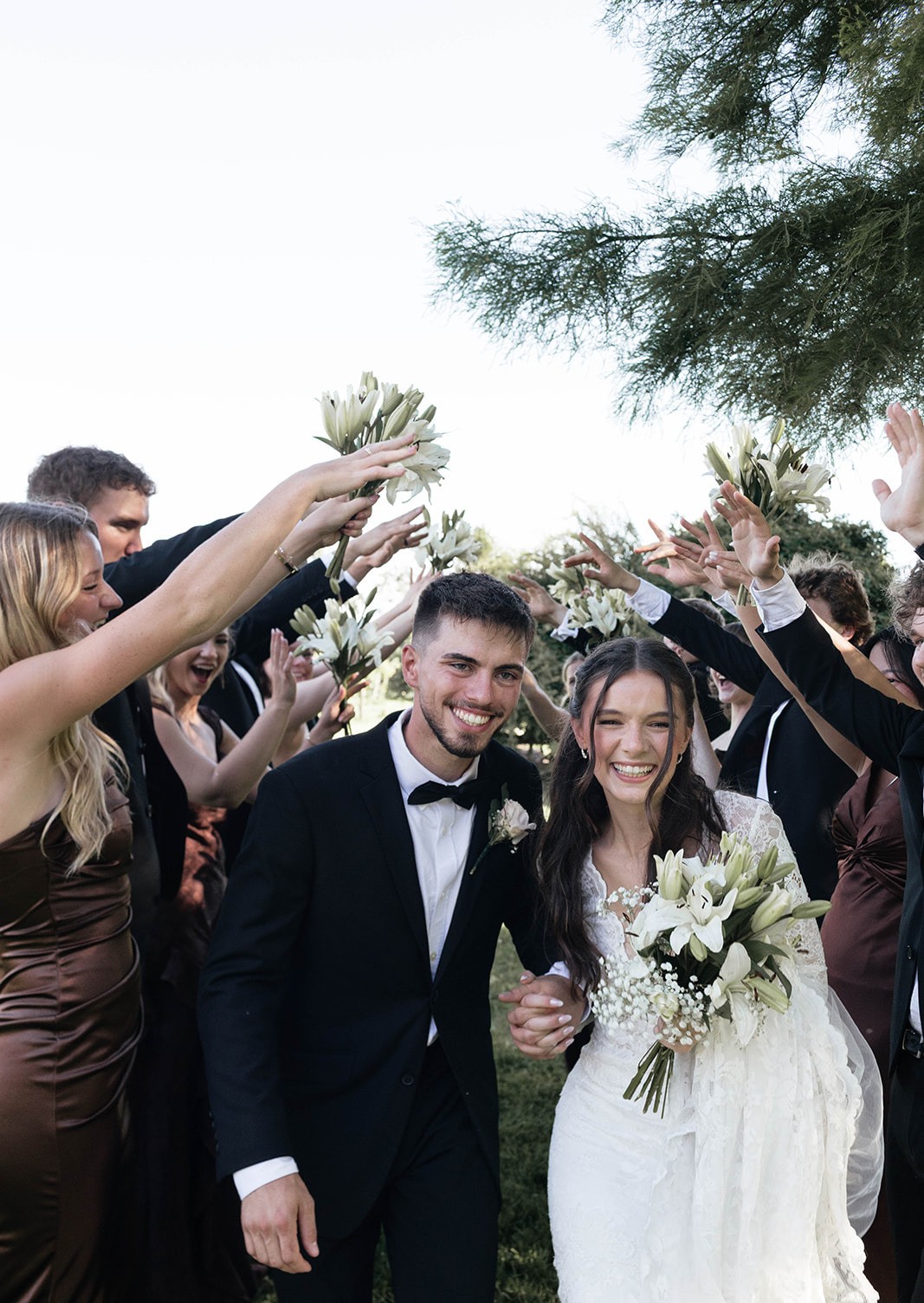 Guests raise white flowers and cheer as the bride and groom walk past laughing