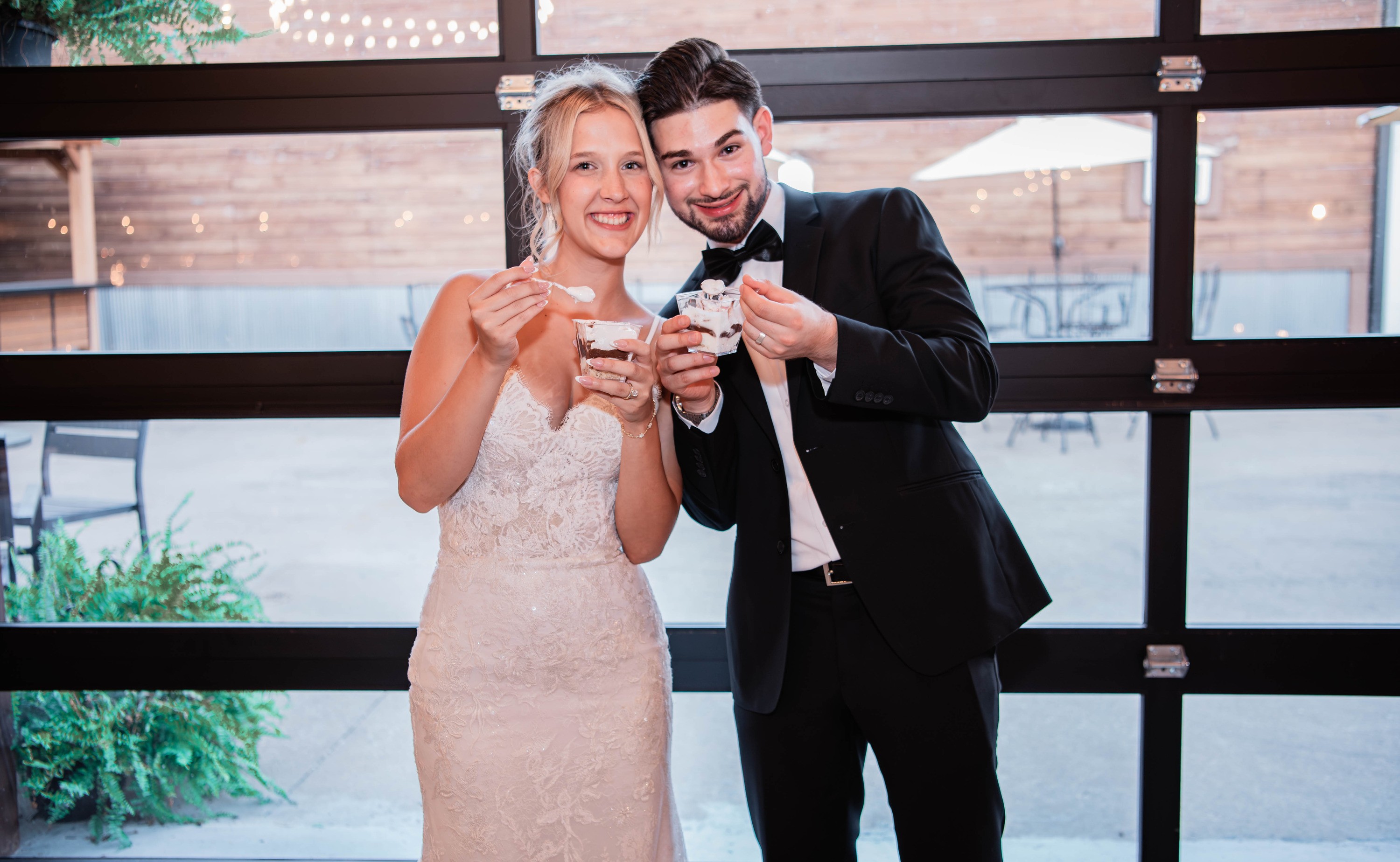 The bride and groom laugh as they eat dessert cups by tall windows