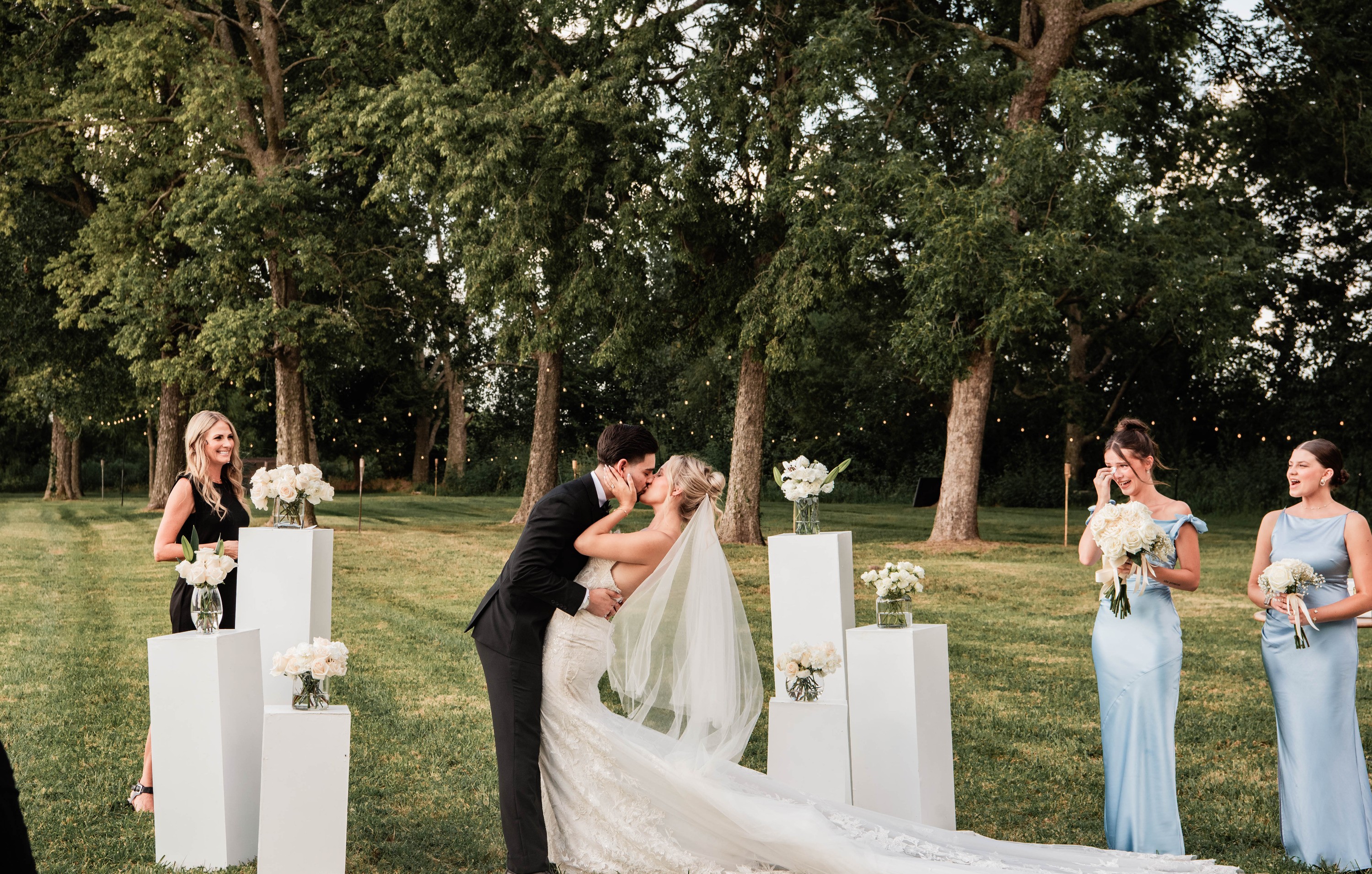 The couple kisses between white pedestals topped with flowers, her long veil on the grass