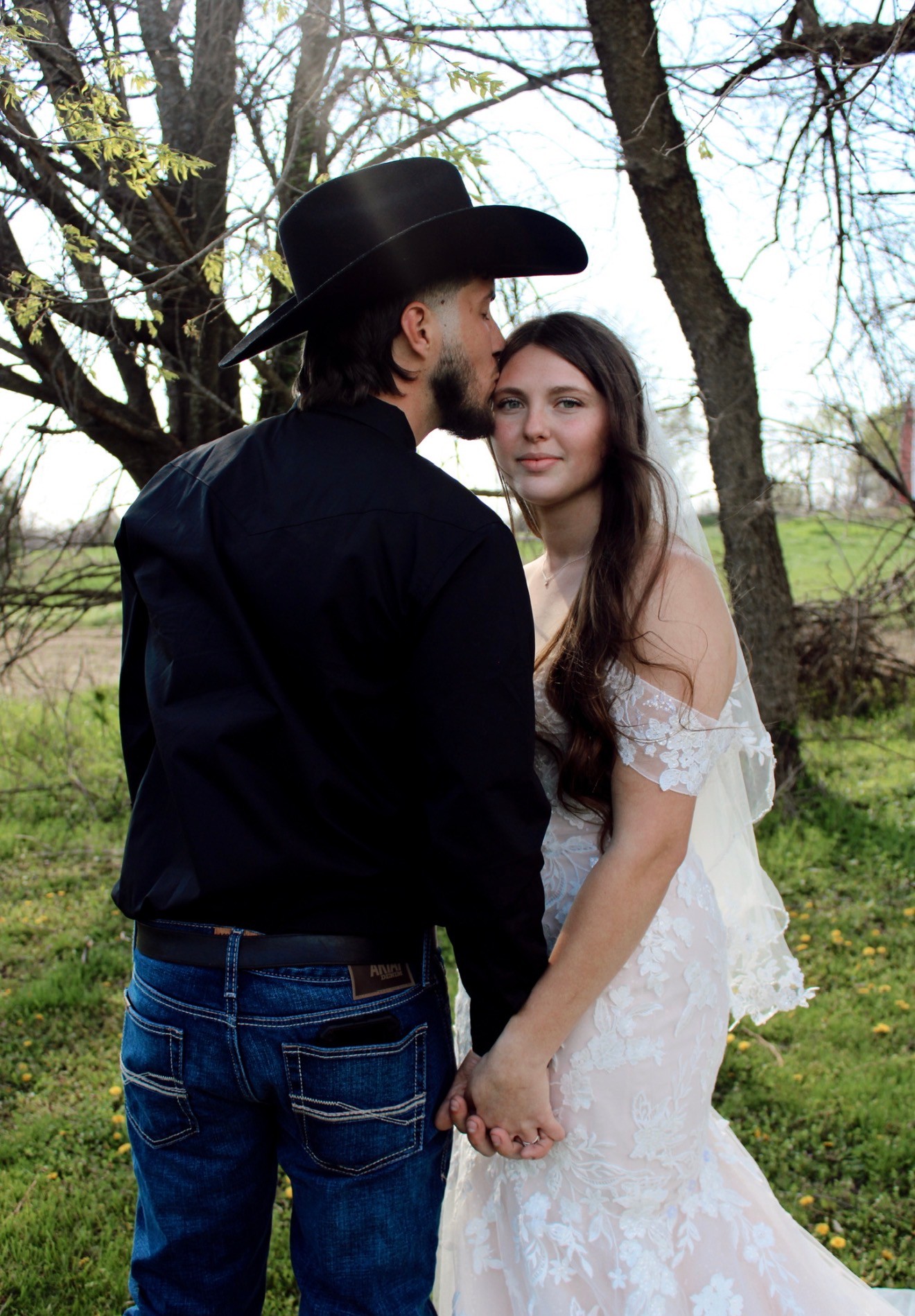 The groom in a black cowboy hat kisses the bride's forehead under the trees