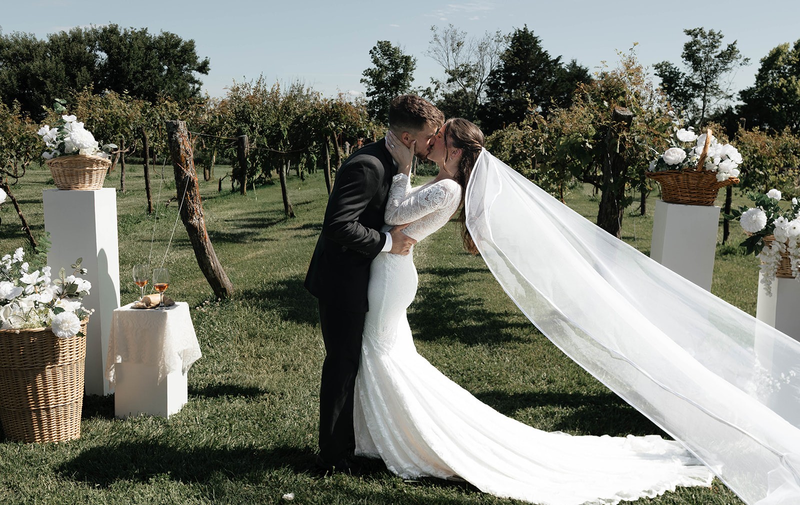 A bride and groom kiss in a green field as her long veil lifts in the wind